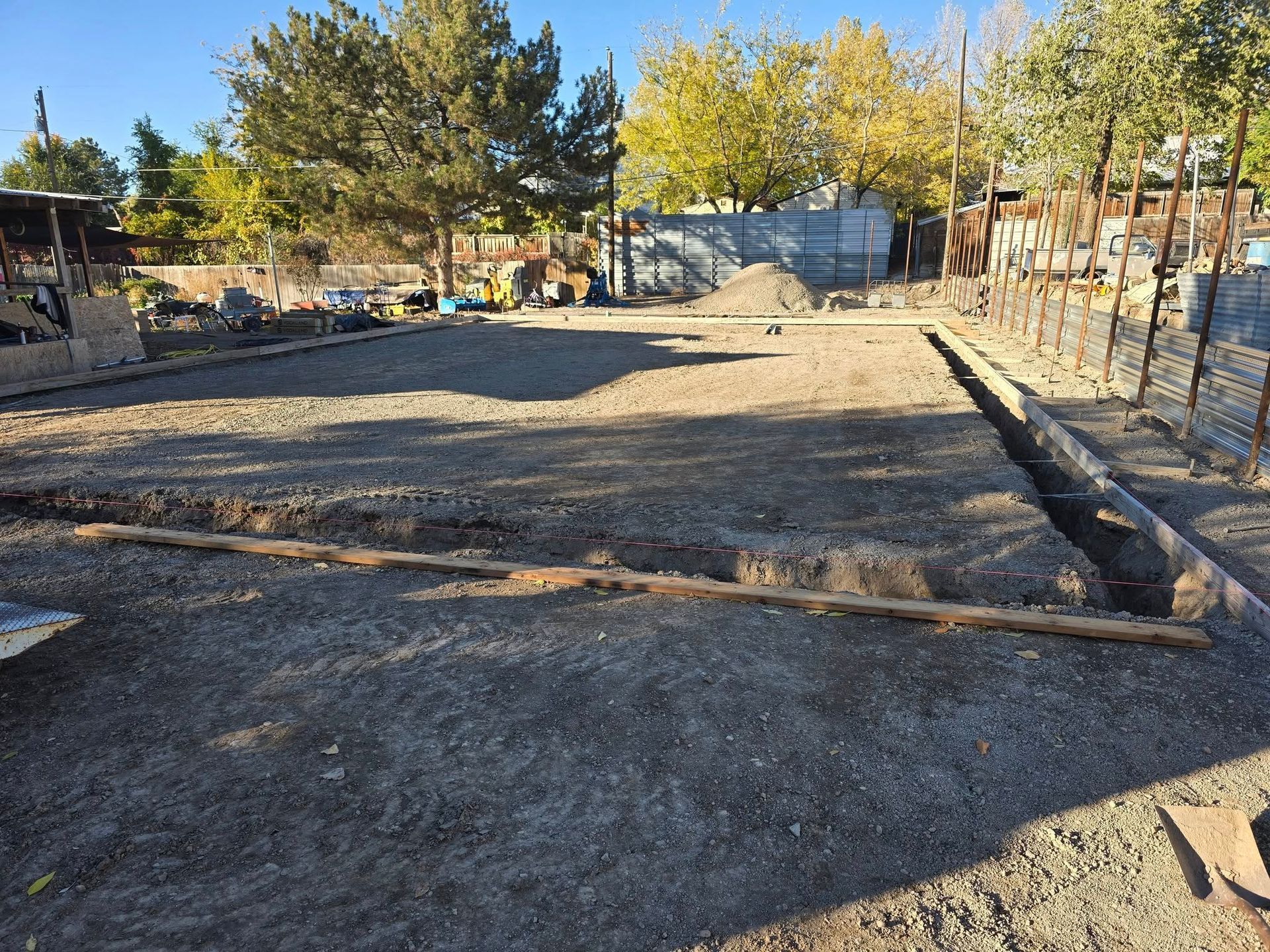 Gravel-filled rectangular construction site, wooden beams defining edges, surrounded by fencing, trees, and a clear blue sky.