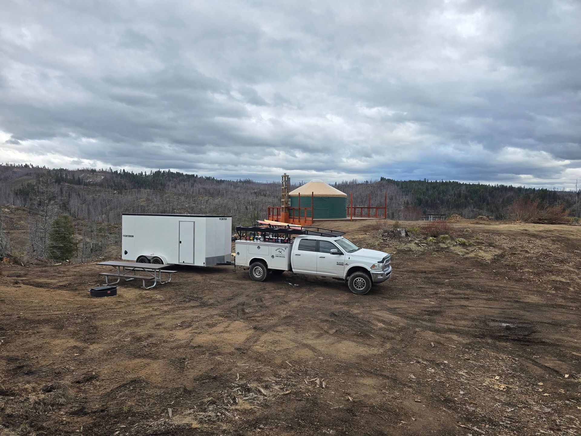 A white pickup truck pulling a trailer on a dirt road, drilling equipment in the background. Overcast sky.