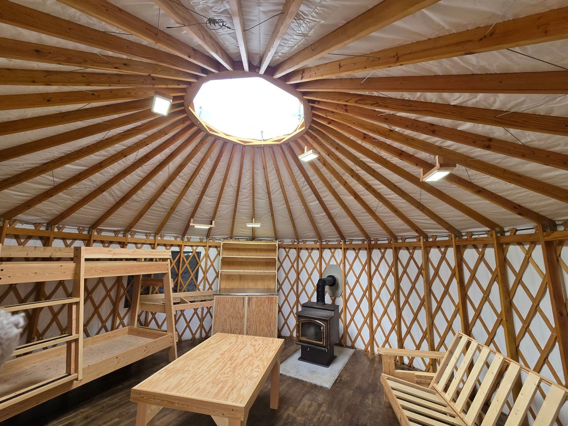 Interior of a yurt with a skylight, wood beams, bunk beds, a table, and a wood-burning stove.