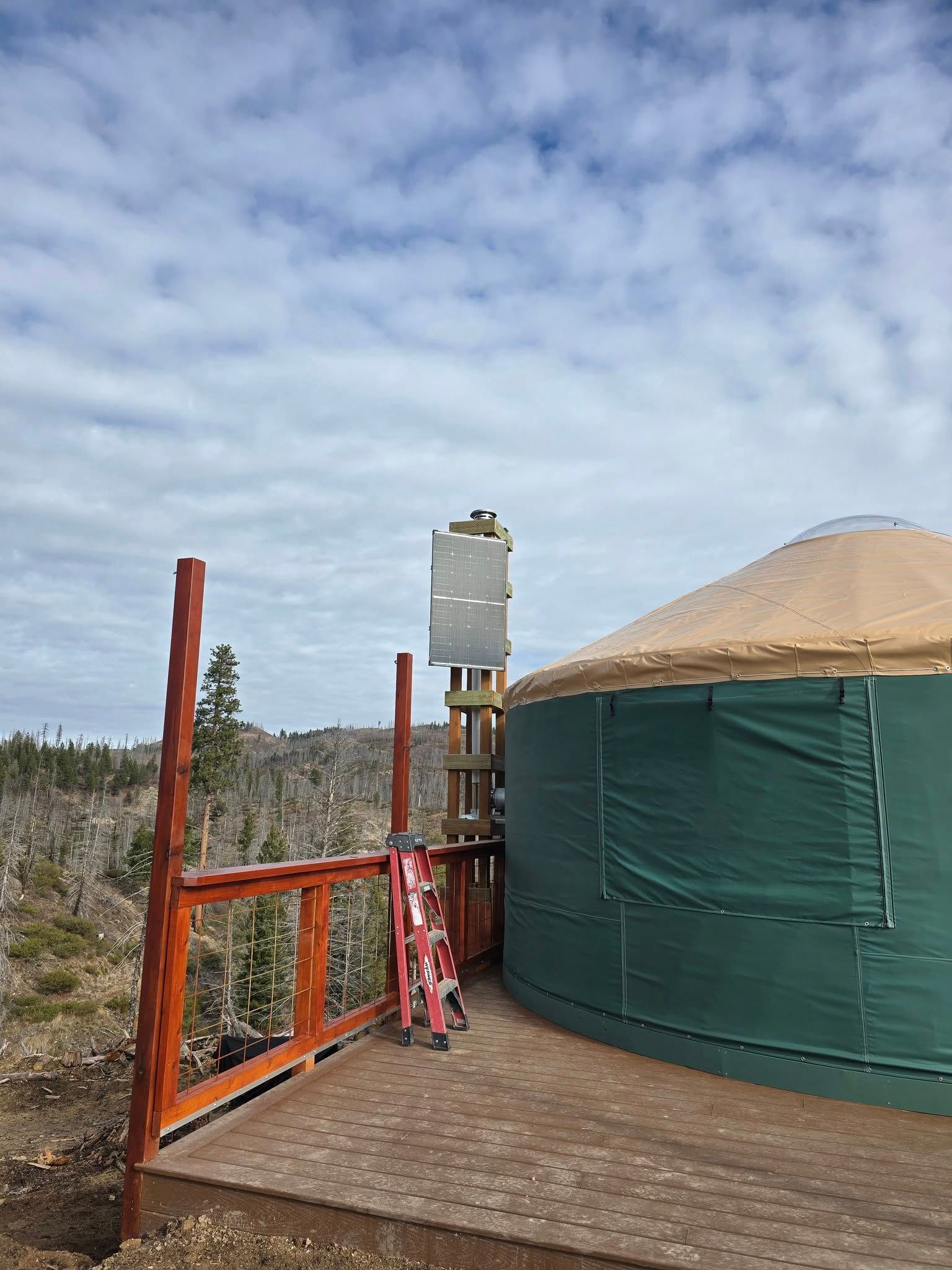 Green yurt with solar panel tower on a wooden deck in a natural setting under a cloudy sky.