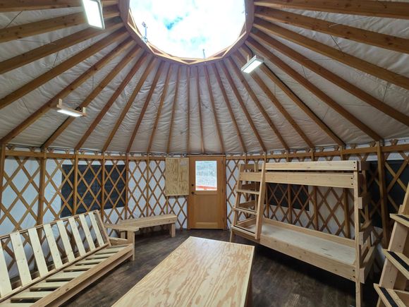 Interior of a yurt with wooden furniture: bunk beds, benches, and a table, with a skylight and wooden lattice walls.