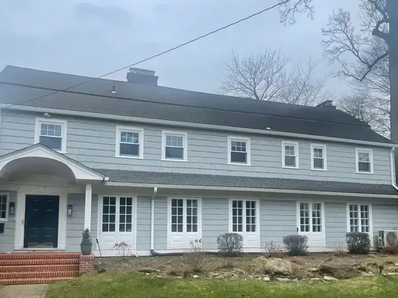 Two-story light blue house with a dark roof and white trim. Gray sky and bare trees.