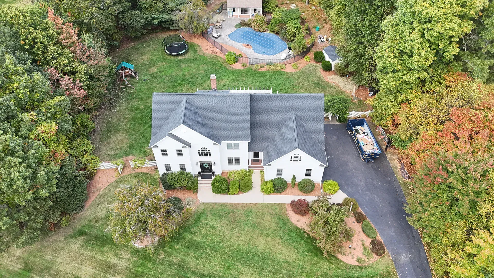 Aerial view of a white two-story house with a dark roof and long driveway surrounded by trees and a pool.
