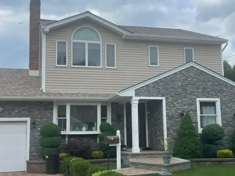 Two-story house with gray stone and beige siding, a brown roof, and a green lawn.