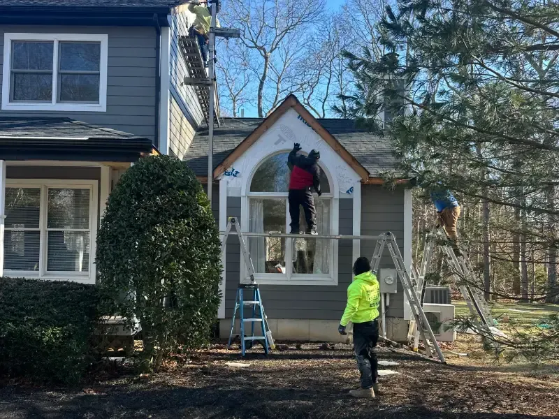 Construction workers on scaffolding, installing siding and roofing on a two-story gray house with large windows; sunny day.