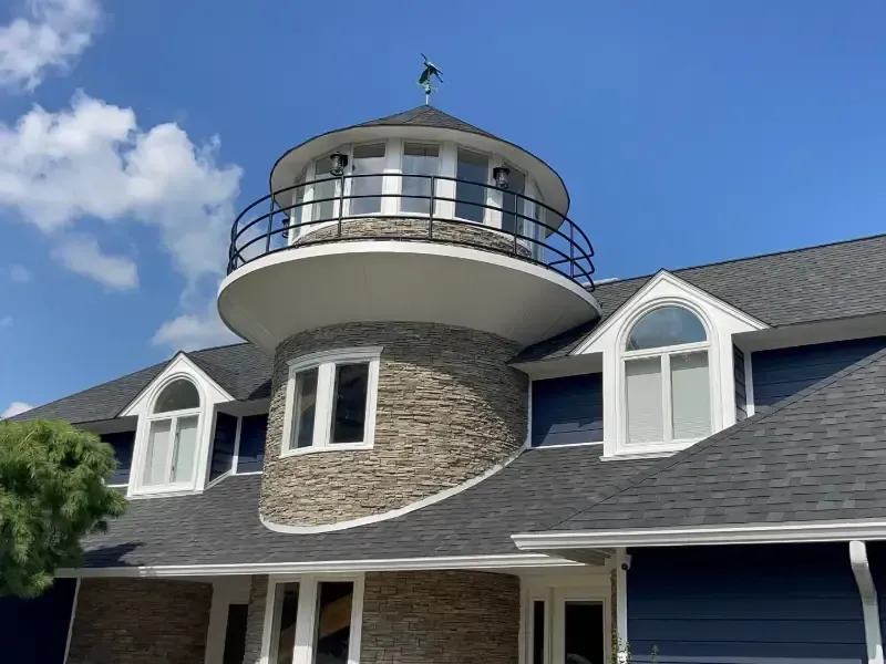 Lighthouse-style building with stone facade, circular tower, and black railing against a blue sky.