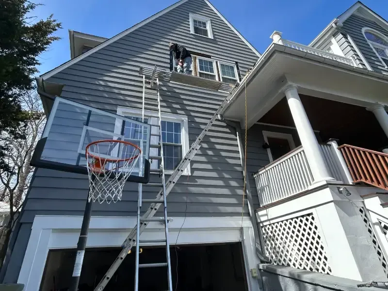 Person on ladder working on a gray house with a basketball hoop in front of a garage.