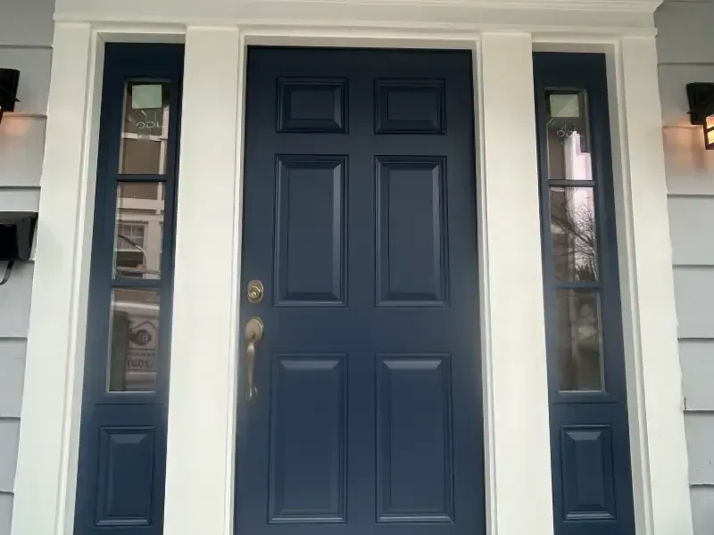 Blue front door with sidelights, white trim, and exterior lights on a gray house.