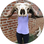 Person holding a ram skull in front of their face. Standing in front of a brick wall.