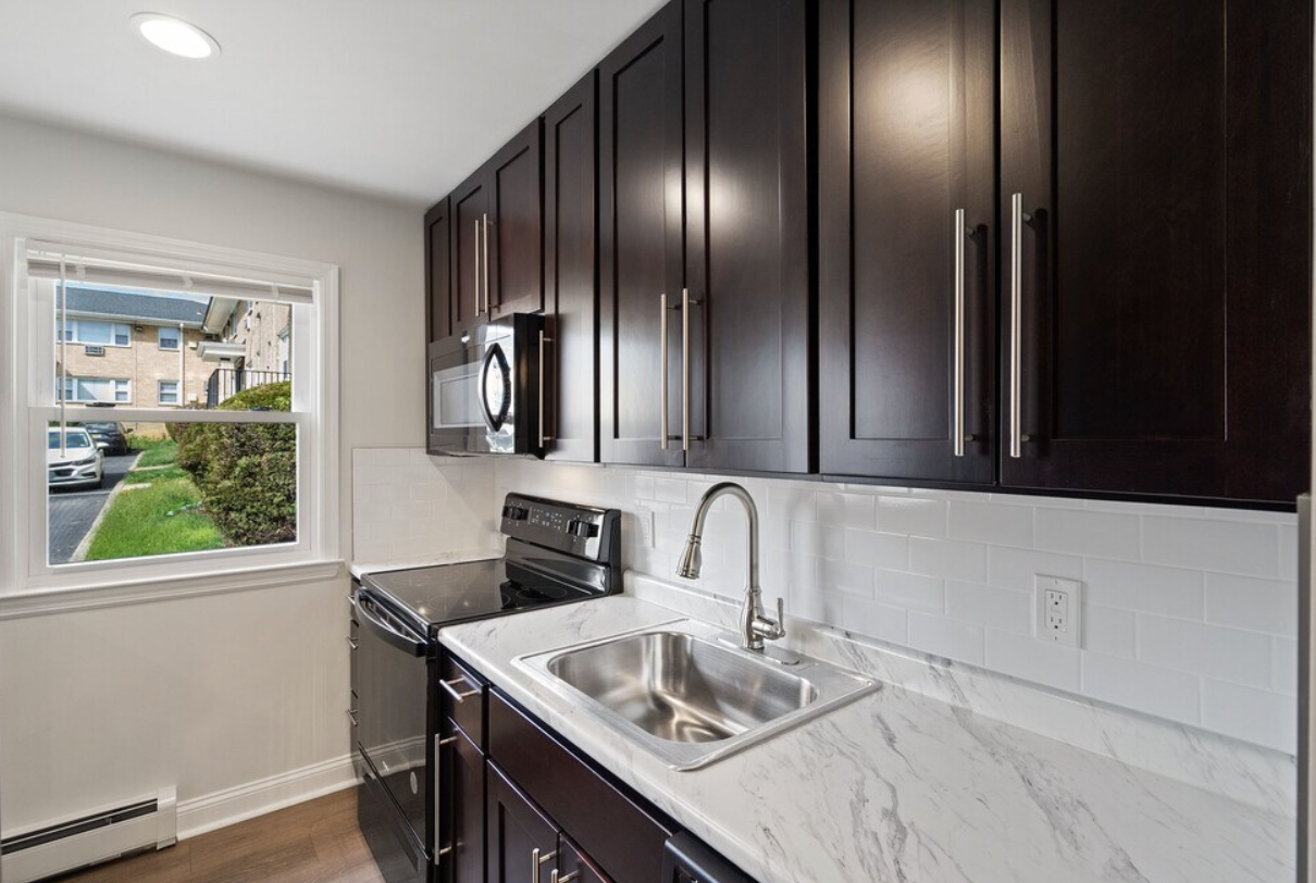 Kitchen with dark cabinets, stainless steel sink, white countertop, and window overlooking a street.
