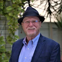 Man with white beard and glasses wearing a black hat and blue blazer, smiling outdoors.