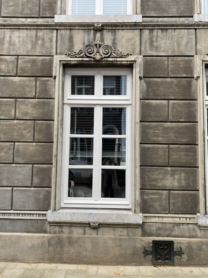 Een hoog raam met een wit kozijn en sierlijst, ingebouwd in een verweerde, grijze stenen gevel.