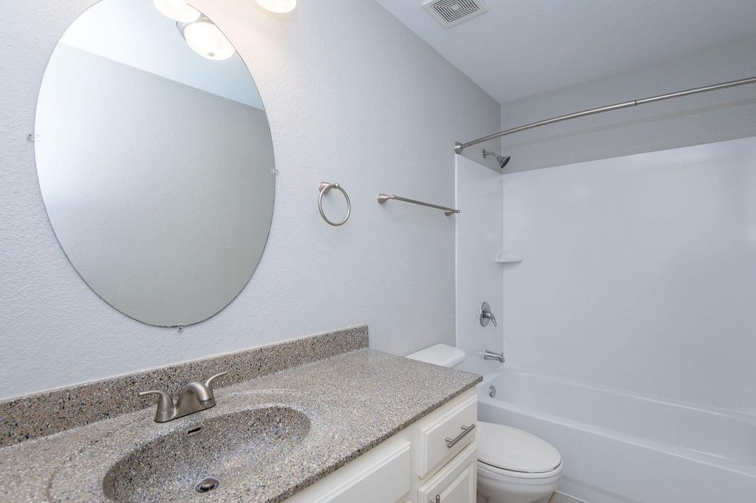 A clean bathroom with a round mirror, gray countertop, white tub, and toilet.