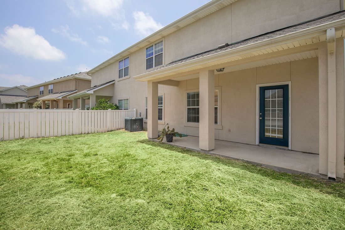 Backyard with green grass, tan house, patio, and white fence on a sunny day.