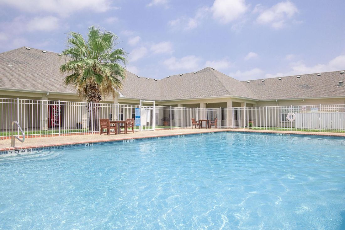 Swimming pool with building and palm tree, fenced in, bright blue water, sunny day.