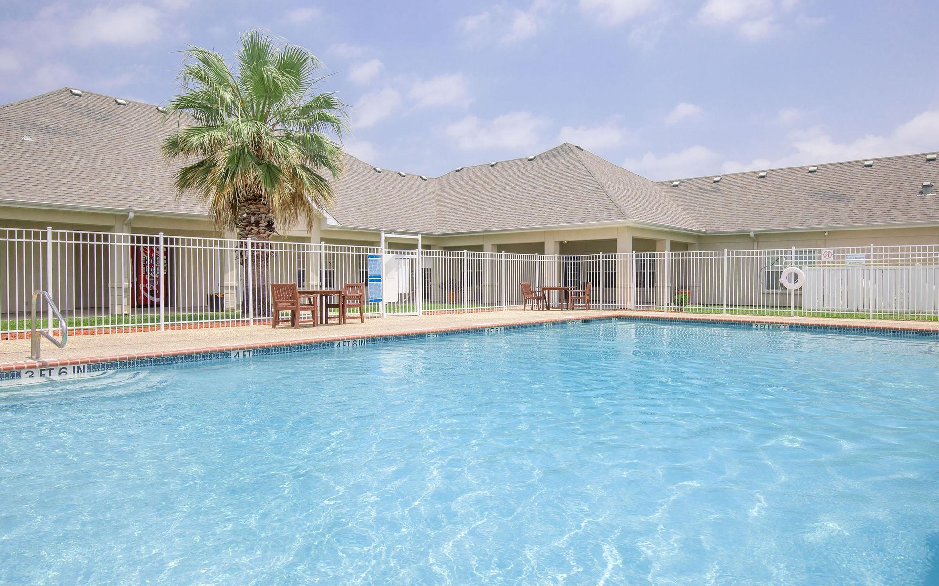 Pool with blue water surrounded by a white fence and building with a palm tree on a sunny day.
