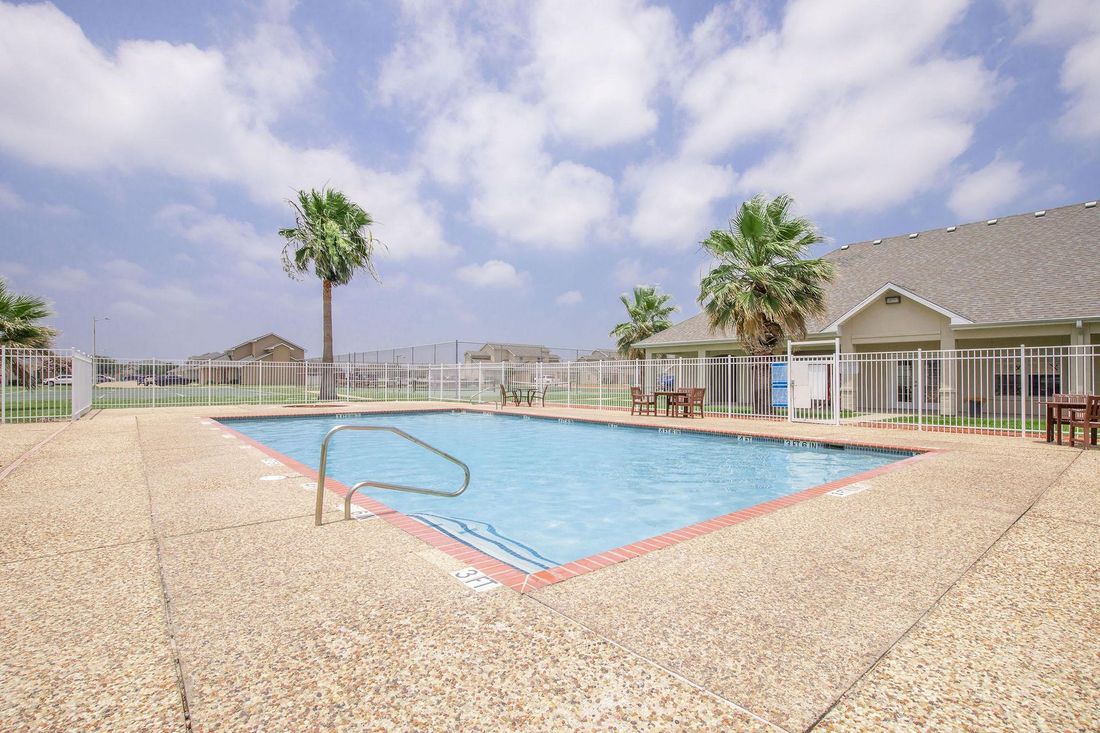 Pool with palm trees, light-colored concrete surround, and a building under a partly cloudy blue sky.
