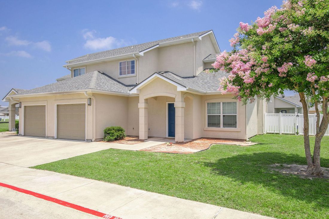 Beige two-story house with attached garage, blue front door, and blooming pink tree on a green lawn.