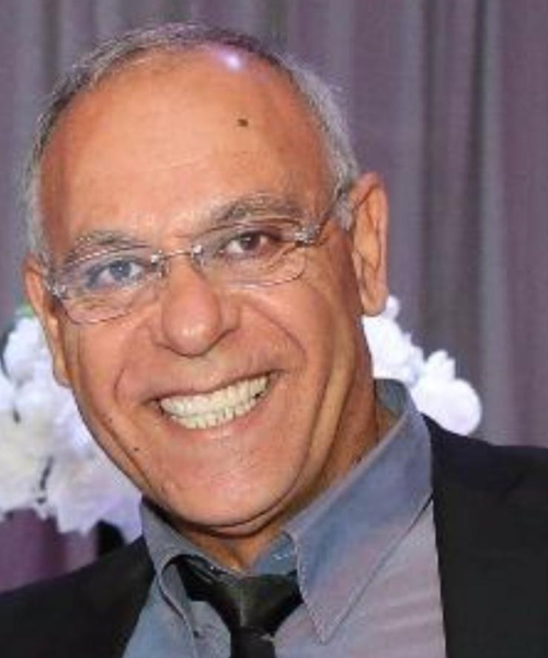 גבר עם משקפיים מחייך, לובש חולצה מכופתרת וז'קט חליפה, בתוך הבית.