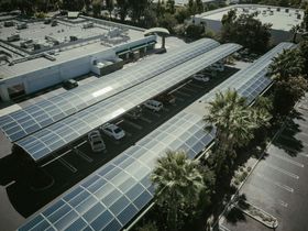 מערכי פאנלים סולאריים המכסים מקומות חניה בחניון. מכוניות חונות מתחת לפאנלים, בניינים ברקע.