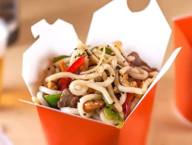 Udon noodles with vegetables and meat in an orange takeout container, sesame seeds sprinkled on top.