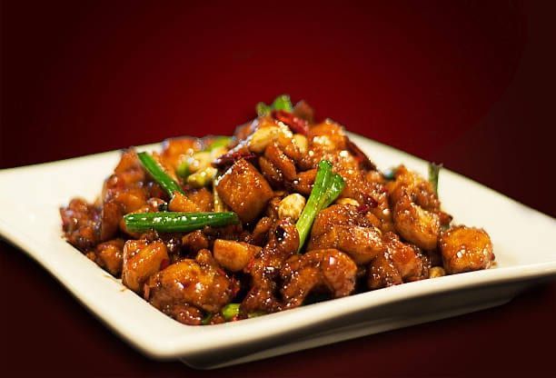 Kung Pao Chicken in a white square dish, featuring chicken pieces, peanuts, chili peppers, and scallions.