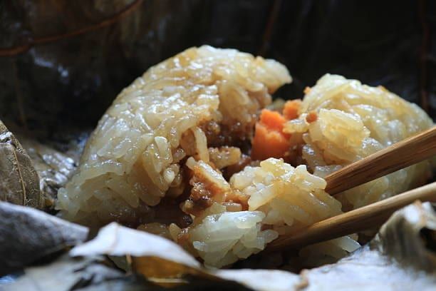 Sticky rice dumpling, partially opened, with meat and carrots, held by chopsticks.