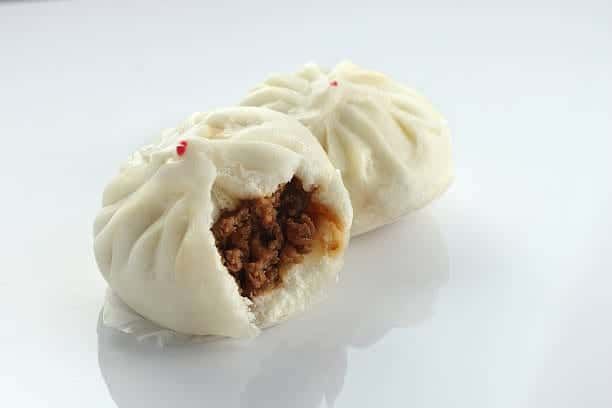 Steamed bao buns, one with a bite taken out, revealing meat filling. White background.