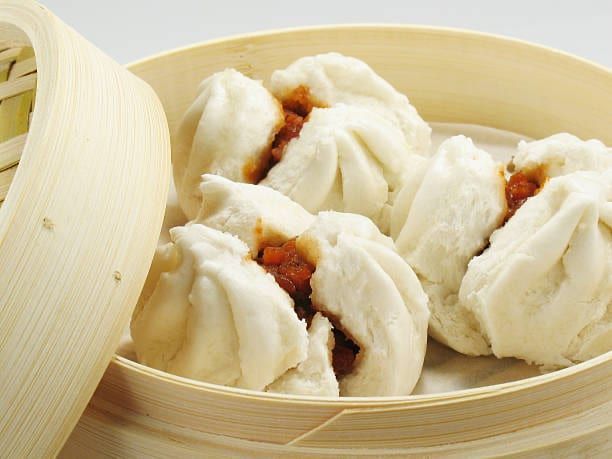Steaming bamboo basket with several fluffy, white bao buns filled with savory meat.