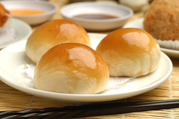 Three golden-brown char siu bao (barbecue pork buns) on a white plate, with chopsticks and dipping sauces nearby.