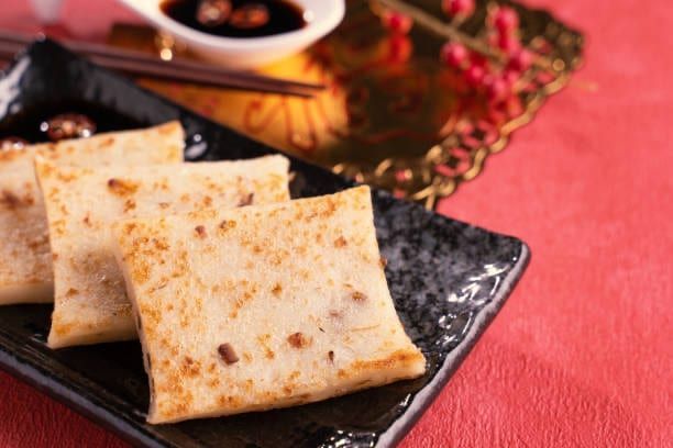 Square turnip cakes on a black plate, served with dipping sauce and chopsticks on a red background.