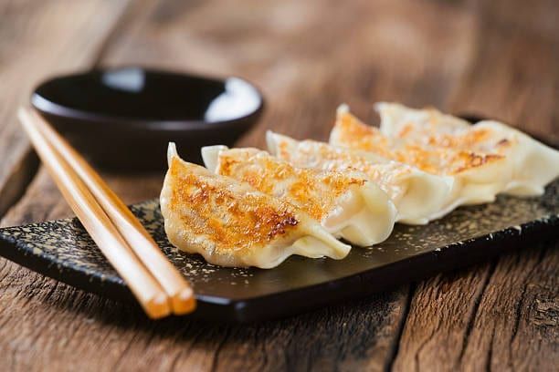 Pan-fried dumplings on a black plate with chopsticks and a small bowl of sauce on a wooden table.