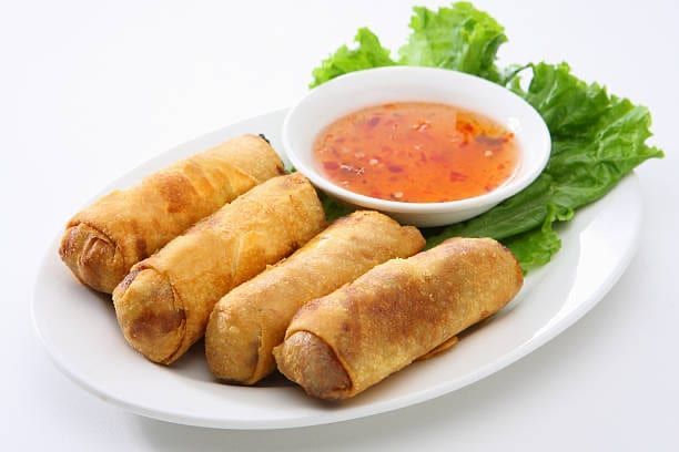 Four golden egg rolls on a plate with a small bowl of dipping sauce and lettuce.