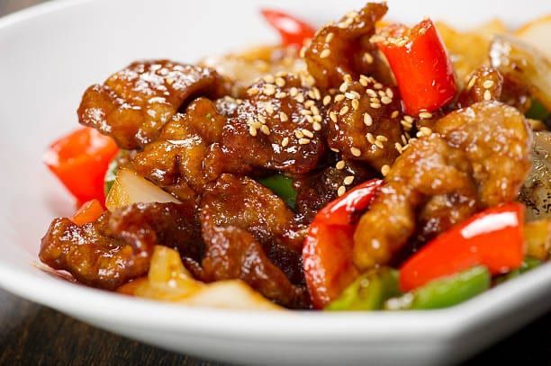 Sweet and sour pork with red bell peppers and sesame seeds in a white bowl.
