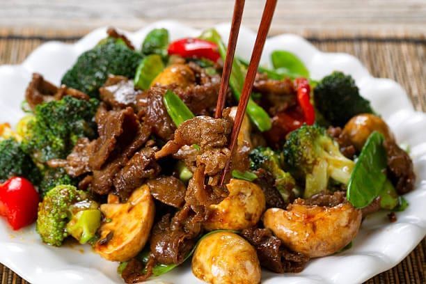 Beef and broccoli stir-fry on a white plate with chopsticks, showcasing cooked mushrooms, bell peppers, and snow peas.