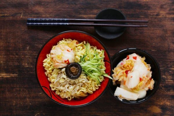 Bowl of noodles with garnishes, side of kimchi, chopsticks on wooden surface.