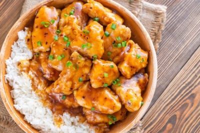 Orange chicken with rice in a wooden bowl, garnished with green onions on a wooden table.
