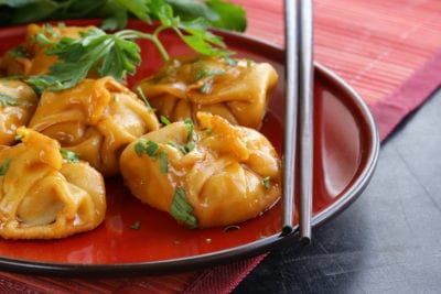 Steamed dumplings in orange sauce on a red plate with chopsticks and parsley garnish.