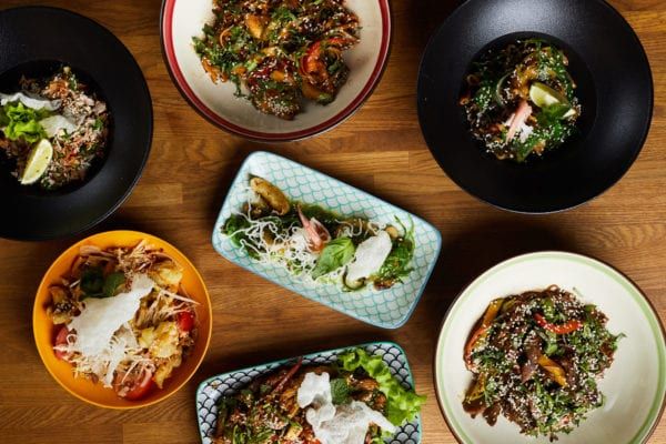 Overhead view of six bowls of colorful Asian dishes on a wooden table.