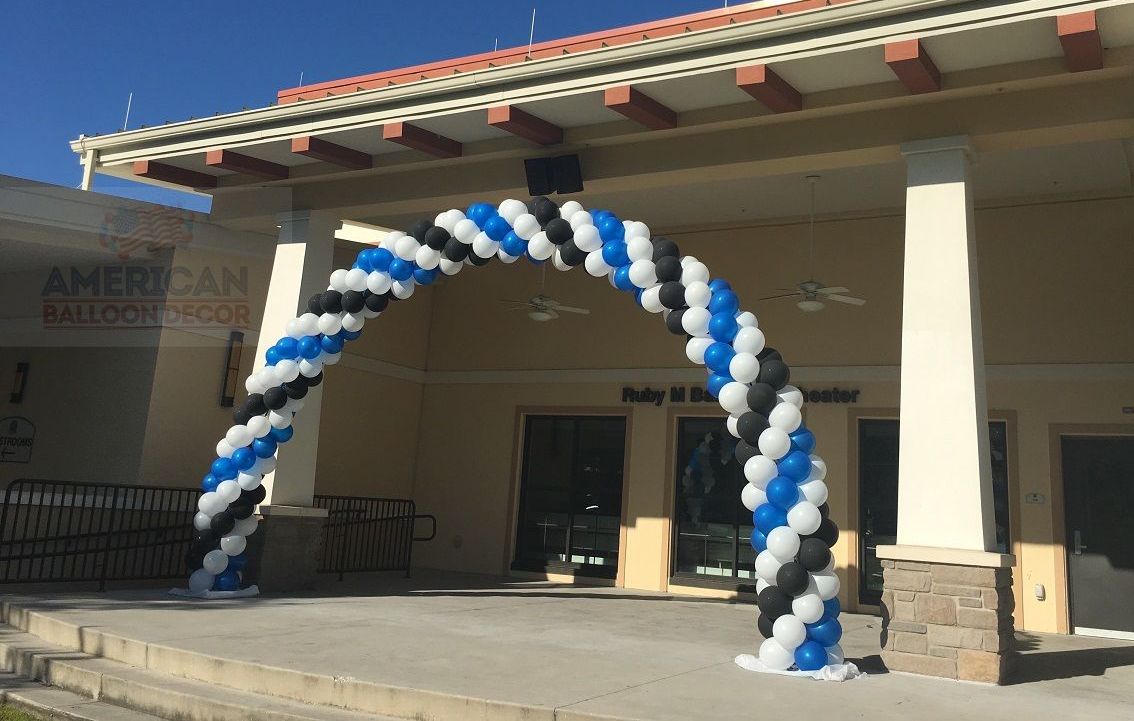 A large arch made of blue , white and black balloons is in front of a building.