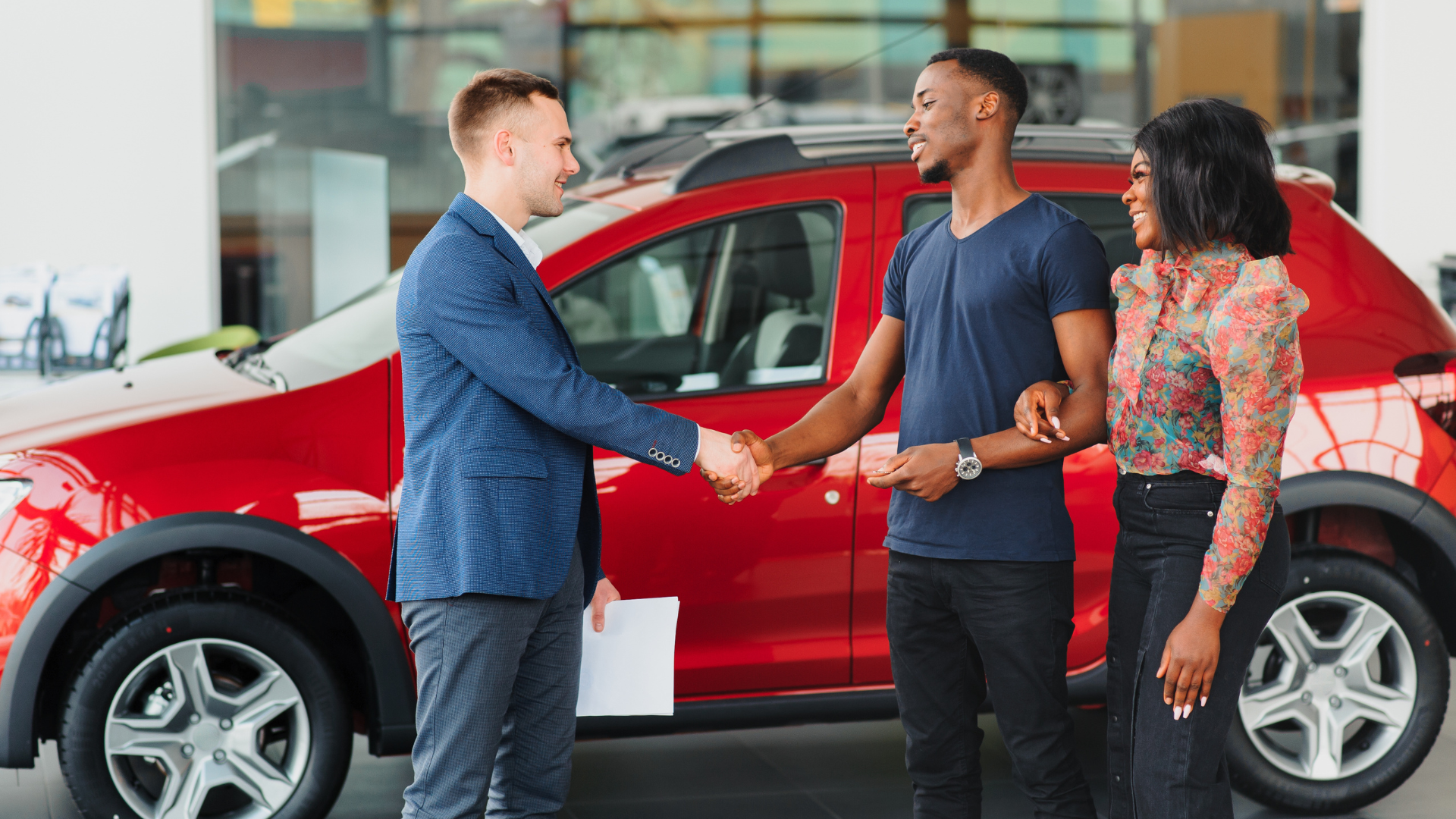A man and a woman are shaking hands in front of a red car.