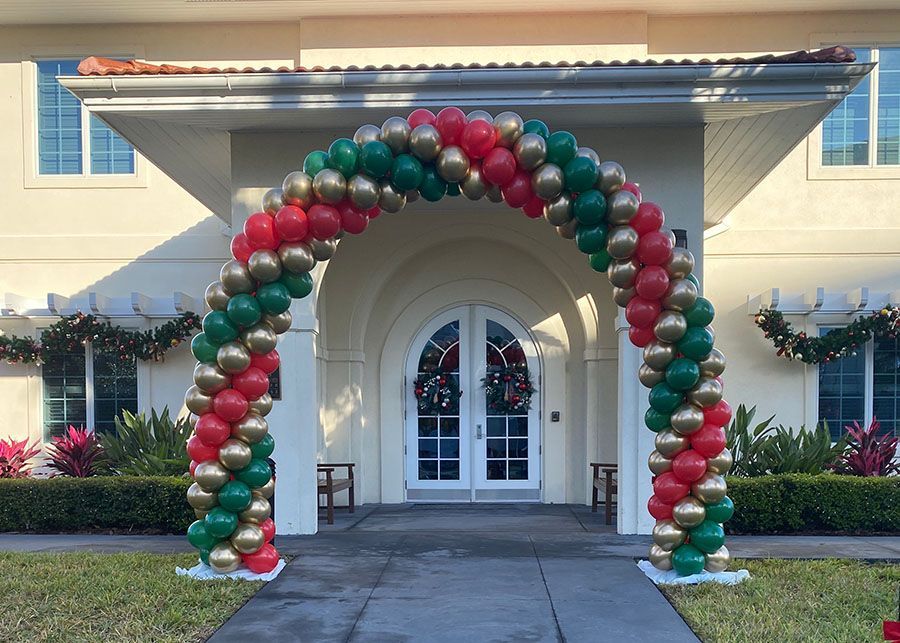 A large arch made of red , green and gold balloons is in front of a house.