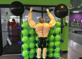 A man made out of balloons is lifting a barbell in a gym.