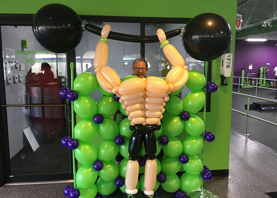 A man made out of balloons is lifting a barbell in a gym.