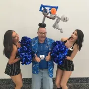 Two cheerleaders are standing next to a man with balloons on his head