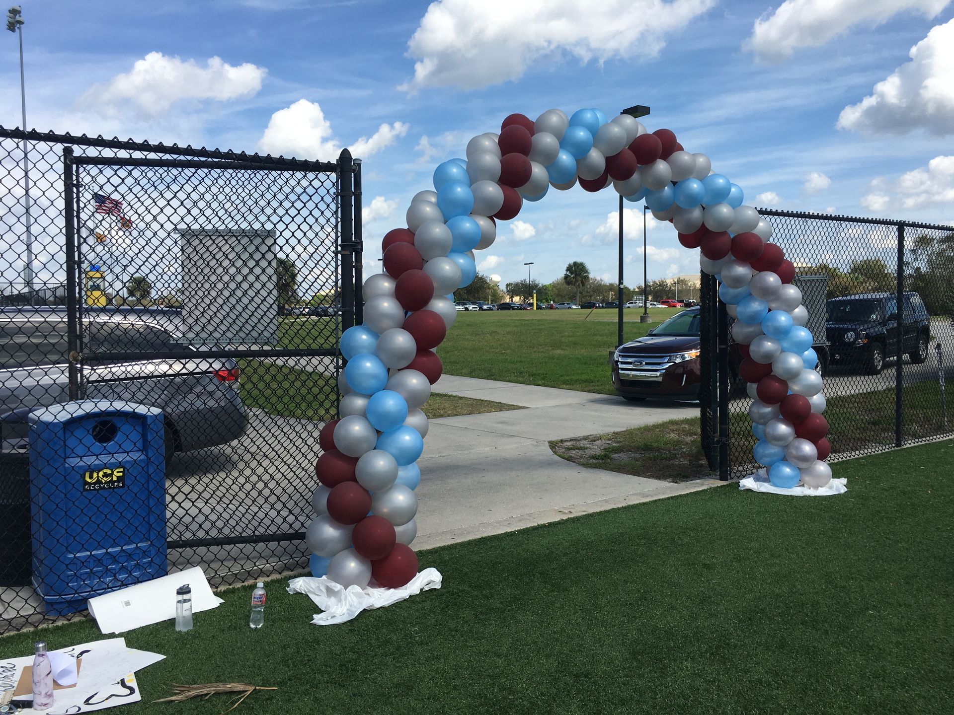 A balloon arch is sitting on the grass in front of a chain link fence.