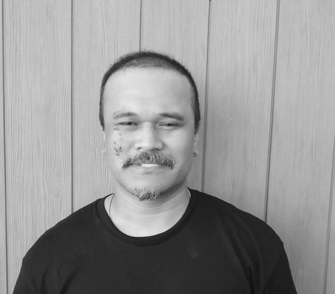 A man with a mustache is wearing a black shirt and standing in front of a wooden wall.