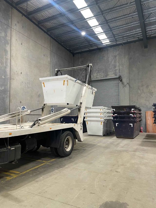 A dumpster truck is parked in a warehouse next to a stack of dumpsters.