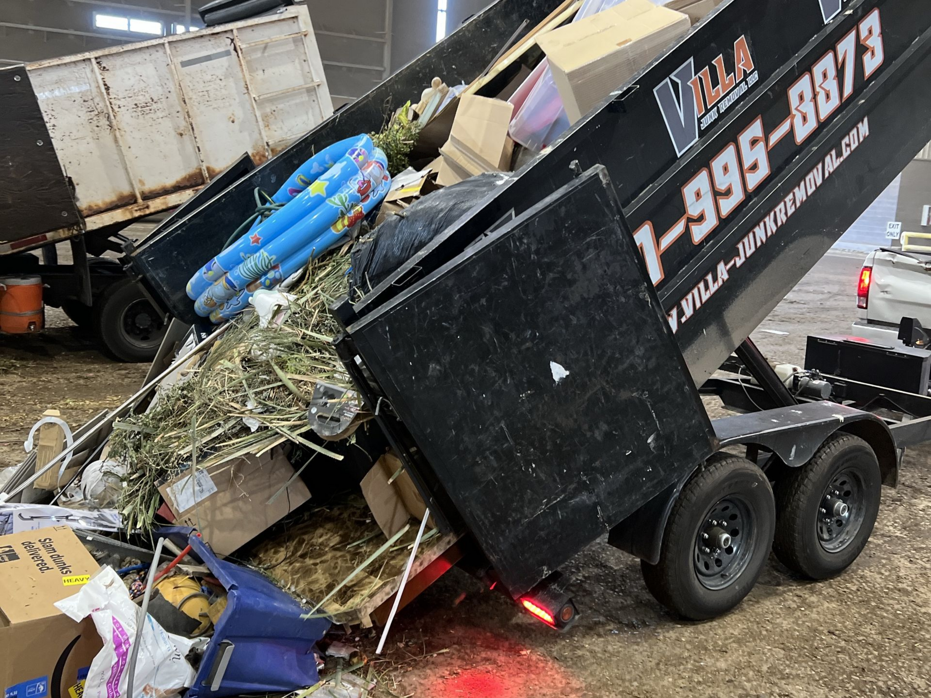Black utility trailer piled high with scrap, debris, and blue containers at a junk yard