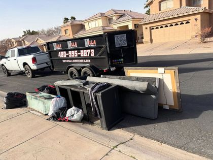 Overturned furniture and trash piled on a sidewalk beside a black utility trailer and white truck in a desert neighborhood